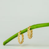 Orecchini a cerchio a treccia in oro giallo appesi a un gambo verde su sfondo chiaro