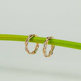 Orecchini a cerchio a treccia in oro tre colori giallo bianco e rosa appessi a un gambo verde su sfondo chiaro